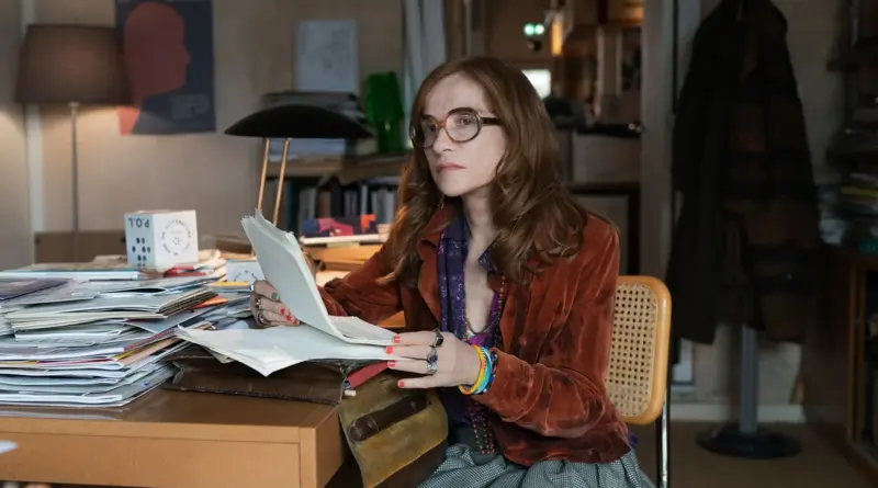 Fotograma de la película Historias Paralelas que muestra a Isabelle Huppert sentada en un escritorio lleno de papeles, mirando fijamente a la cámara.