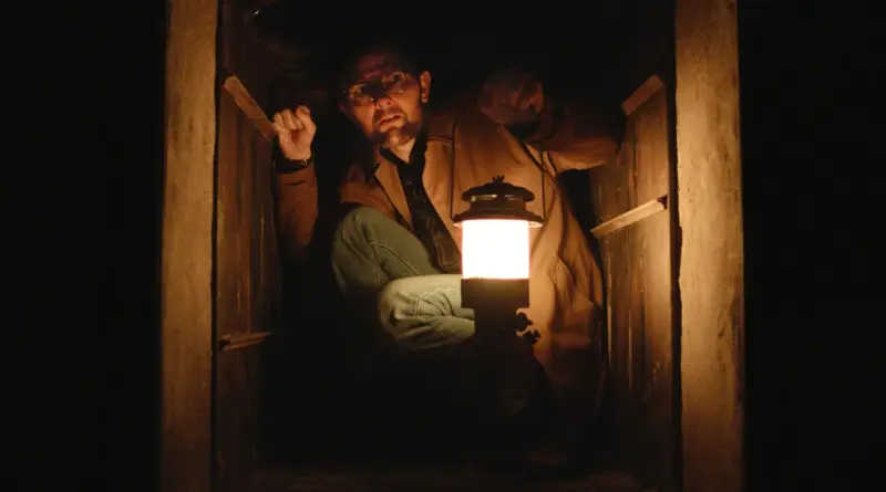 Adam Scott agachado en un espacio pequeño de madera, iluminando la oscuridad con una linterna de gas, en 'Hokum'.