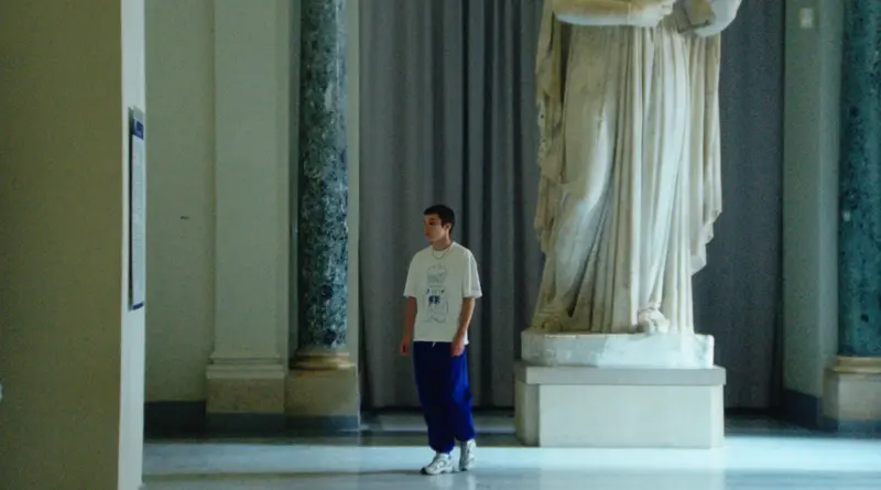 Fotograma de la película La Gradiva (2026) con un joven caminando frente a una gran estatua clásica en un museo.