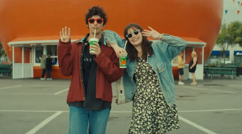 Fotograma de la película La ironía del amor (Mile End Kicks), con una pareja sonriente con gafas de sol tomando batidos frente a una gran estructura naranja.