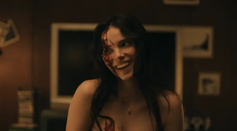 r plano de una mujer joven sonriendo intensamente con sangre en el rostro y el pecho, escena de la película Obsession.