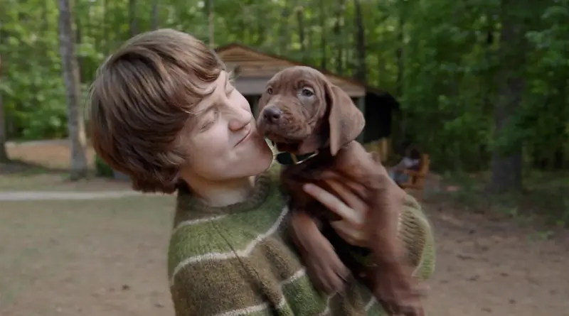 El joven protagonista de Scooby-Doo: El origen sonríe con ternura mientras sostiene en brazos al pequeño cachorro marrón en un entorno natural.