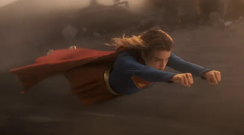 Supergirl vuela horizontalmente a gran velocidad con los puños al frente, vistiendo su traje azul y capa roja, en un entorno envuelto en polvo y niebla.