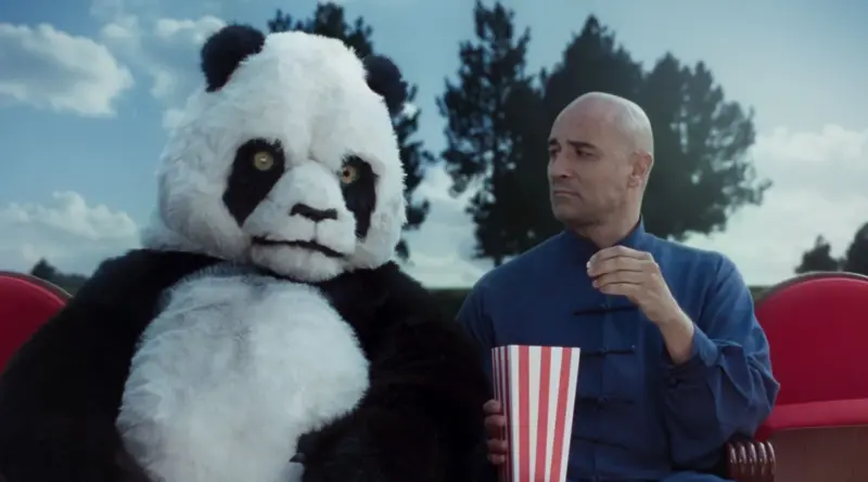 Un hombre vestido de azul mira con desconfianza a una persona disfrazada de oso panda gigante mientras comen palomitas, en Una pistola, una bala y un oso panda.