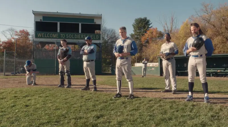 Escena de la película Eephus donde varios jugadores del equipo Riverdogs están de pie en silencio en un campo de béisbol otoñal frente a un antiguo marcador.
