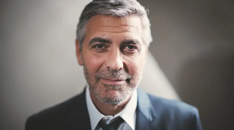 Retrato en primer plano de George Clooney sonriendo levemente, vistiendo un traje oscuro, camisa blanca y corbata negra.