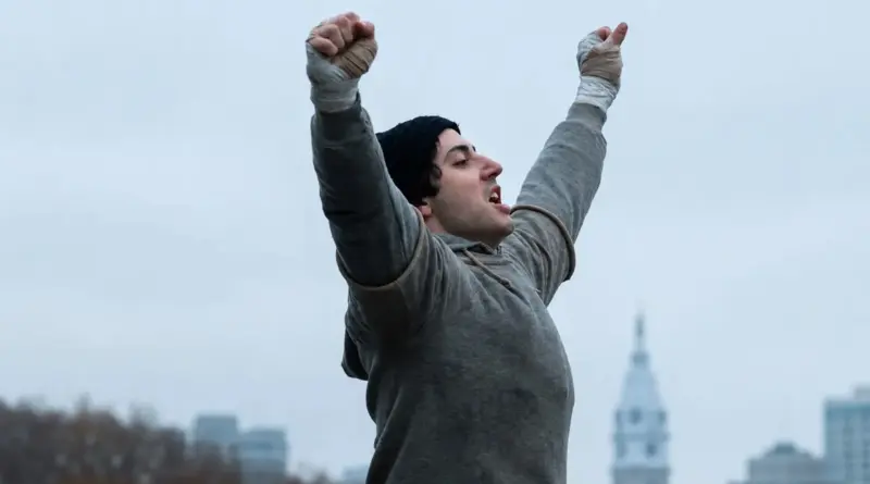 Hombre en sudadera gris y gorro negro alzando los puños vendados hacia el cielo en señal de triunfo, de la película 'Yo soy Rocky' ('I Play Rocky').