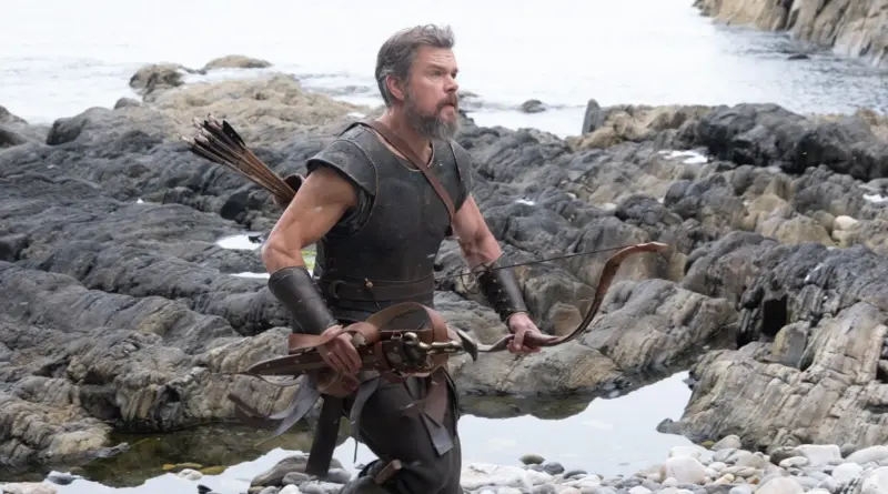 Escena de la película La Odisea donde un guerrero barbudo con armadura antigua camina por una costa rocosa empuñando un arco.