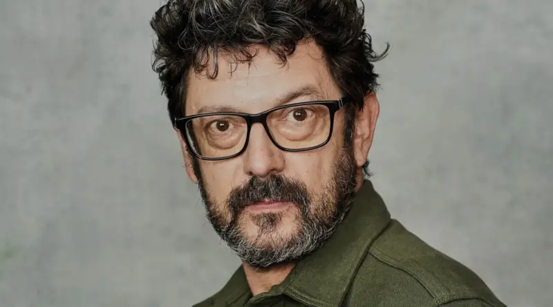 Primer plano del actor Manolo Solo sobre un fondo gris, llevando gafas de pasta negra y una camisa verde oliva.