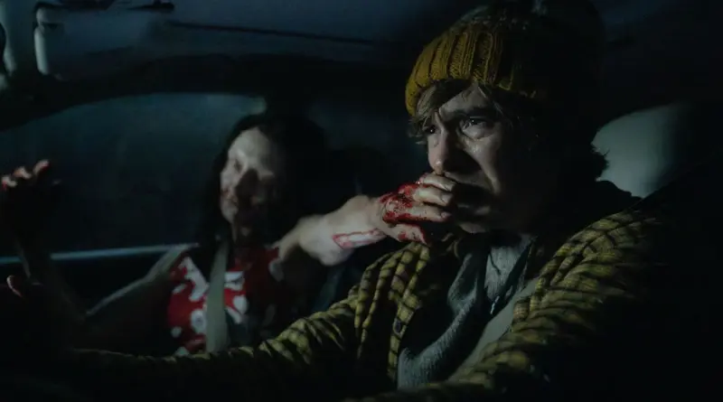 Austin Abrams, visiblemente aterrorizado, se tapa la boca dentro de un coche oscuro mientras una mujer zombi lo ataca en Resident Evil.