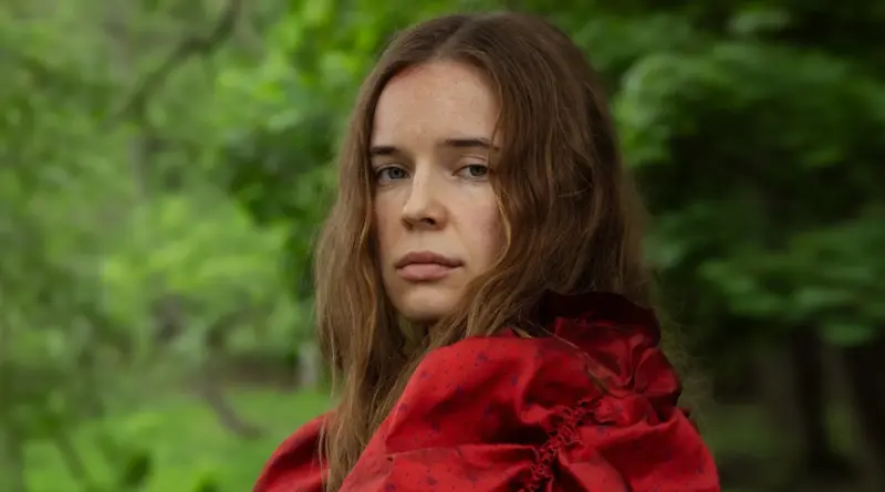 Primer plano de la protagonista de Tell Everyone con expresión seria, vistiendo un vestido rojo en un bosque verde.