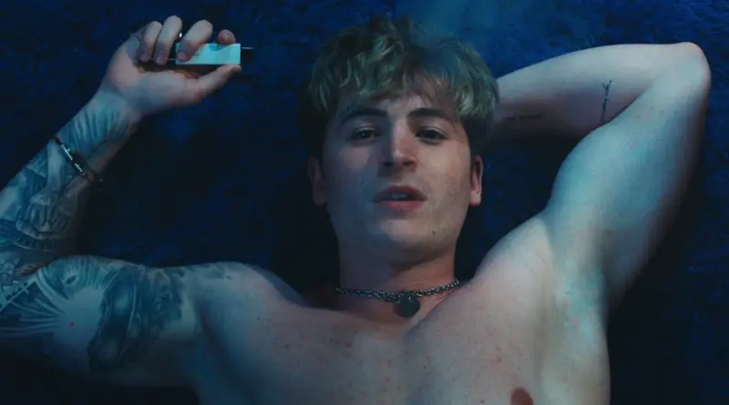Primer plano de un joven tatuado de la película Blue Film tumbado sobre una superficie de pelo azul, sosteniendo un pequeño objeto blanco.