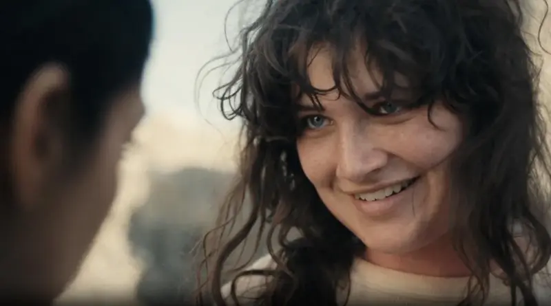 Primer plano de una mujer de cabello rizado oscuro y ojos claros mirando fijamente hacia un lado con una leve sonrisa.