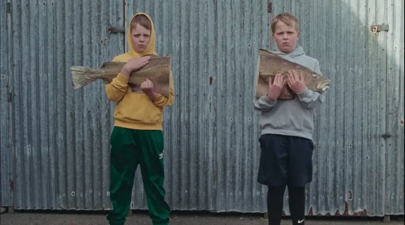 Dos niños de la película El amor que permanece de pie, muy serios, frente a una pared de chapa metálica ondulada; cada uno sostiene una gran mitad de un enorme pez cortado.