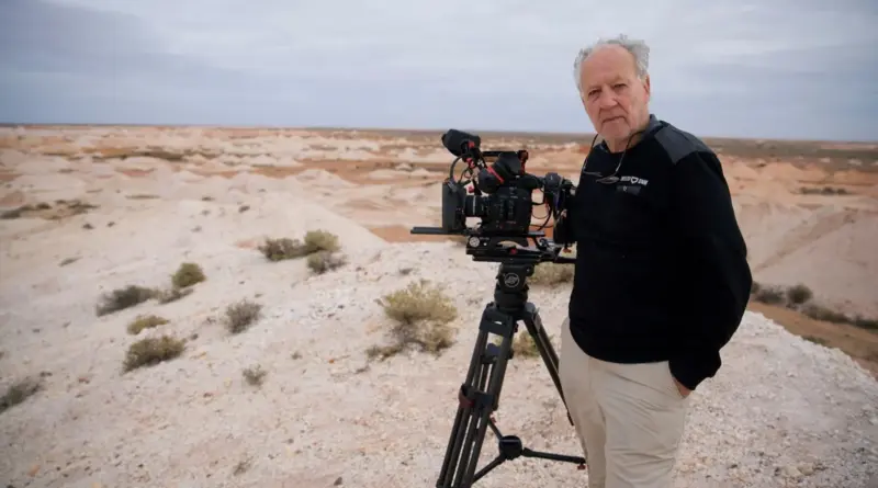 El director Werner Herzog de pie junto a una cámara de cine profesional en un vasto paisaje desértico.