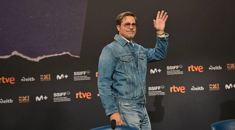 Brad Pitt saludando con la mano en alto frente a un panel oscuro del Festival de San Sebastián.