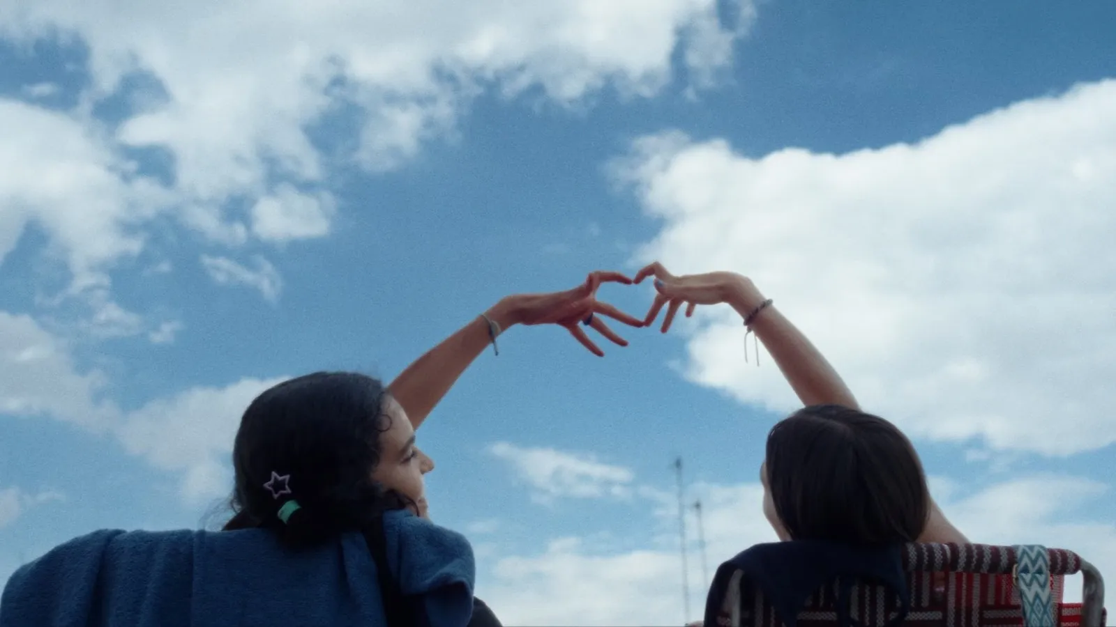 Las protagonistas de Chicas tristes forman un corazón en el aire uniendo sus manos frente a un luminoso cielo azul con nubes.