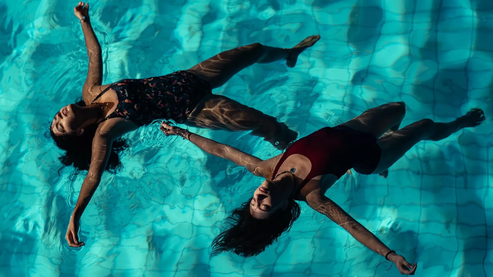 Las dos amigas protagonistas de la película Chicas tristes flotando relajadas bocarriba en el agua cristalina de una piscina iluminada por el sol.