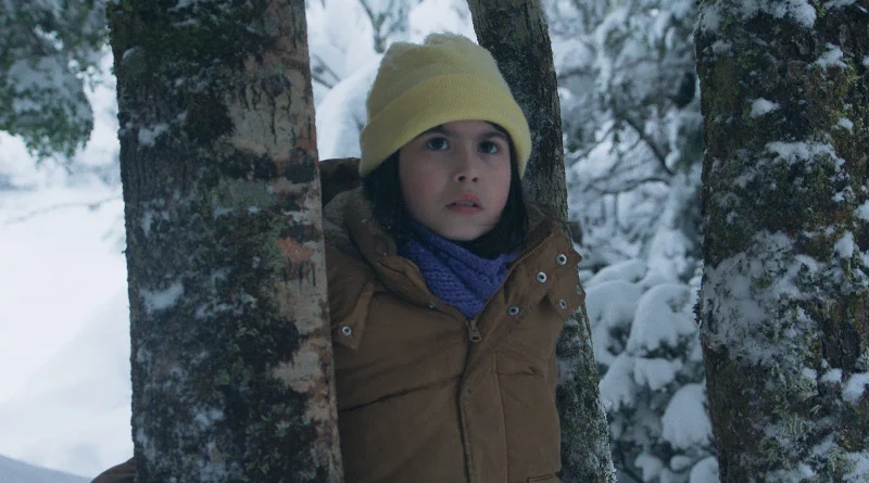 Una niña pequeña se asoma con cautela entre dos árboles nevados en el bosque en una imagen de la película El deshielo.