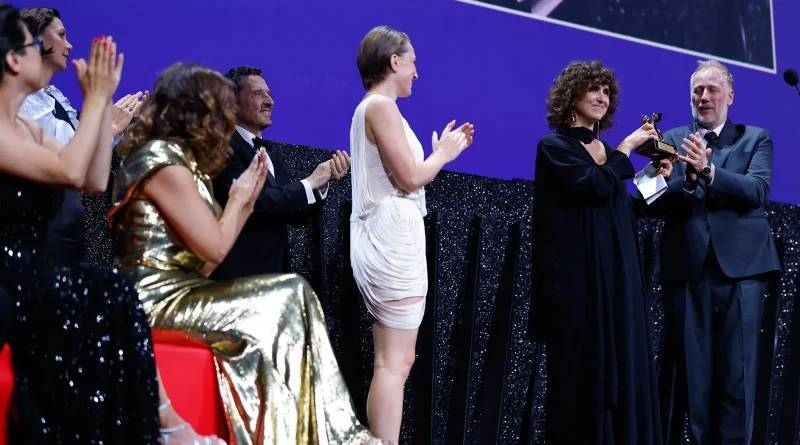 Mujer con vestido negro sonríe mientras sostiene el trofeo del León de Oro en un escenario, rodeada de asistentes que le aplauden en el Festival de Venecia 2026.