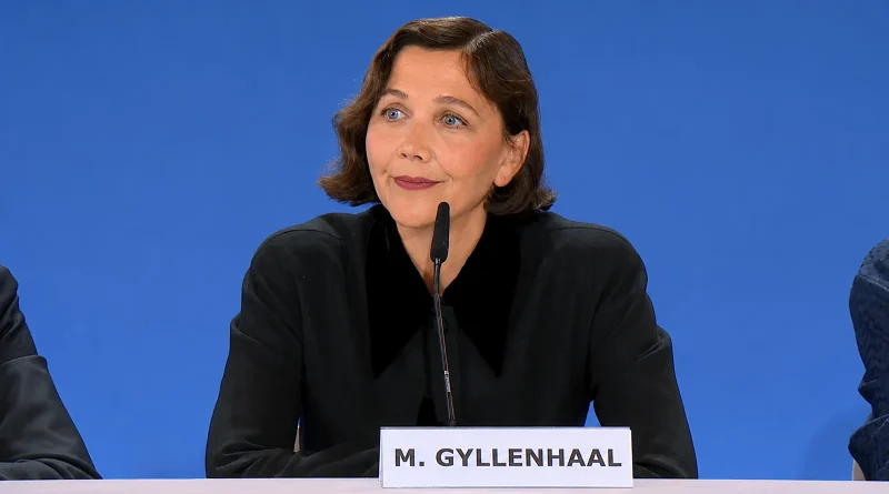 Fotografía de busto de Maggie Gyllenhaal esbozando una leve sonrisa irónica durante una rueda de prensa. Viste una sobria blusa negra con un amplio cuello de terciopelo. Delante de ella hay un micrófono y un letrero blanco con el texto "M. GYLLENHAAL", todo ello recortado sobre un fondo liso de color azul vibrante.