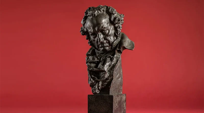 Estatuilla de bronce de los Premios Goya que representa el busto del pintor Francisco de Goya sobre un fondo rojo uniforme.