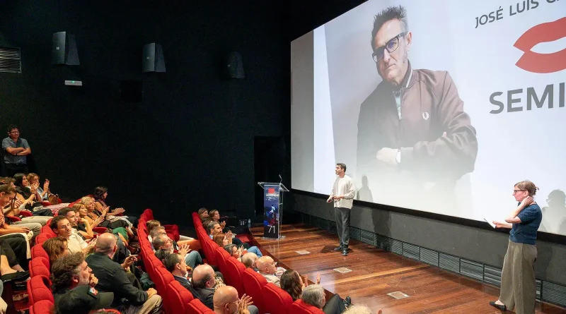 Fotografía de una presentación en una sala de cine durante la Seminci, donde dos ponentes en un escenario se dirigen a un público que aplaude frente a una gran pantalla con el retrato y nombre de José Luis Cienfuegos.