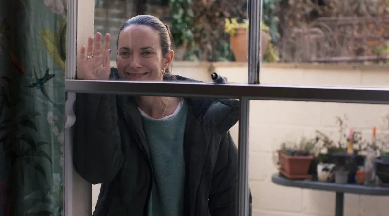 La protagonista de Tender Loving Care asomándose y saludando con la mano a través del cristal de una ventana desde el exterior de una casa.