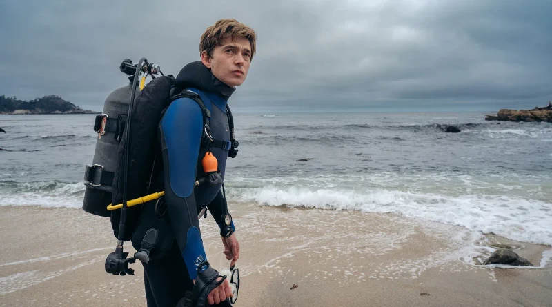 El protagonista de Whalefall, Austin Abrams, en la orilla de una playa nublada, cargando su equipo de buceo antes de entrar al mar.