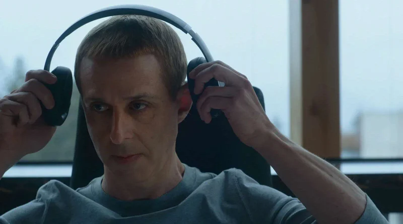 Fotograma de la película El precio de la red en el que aparece un hombre con el pelo muy corto poniéndose unos auriculares negros de diadema.