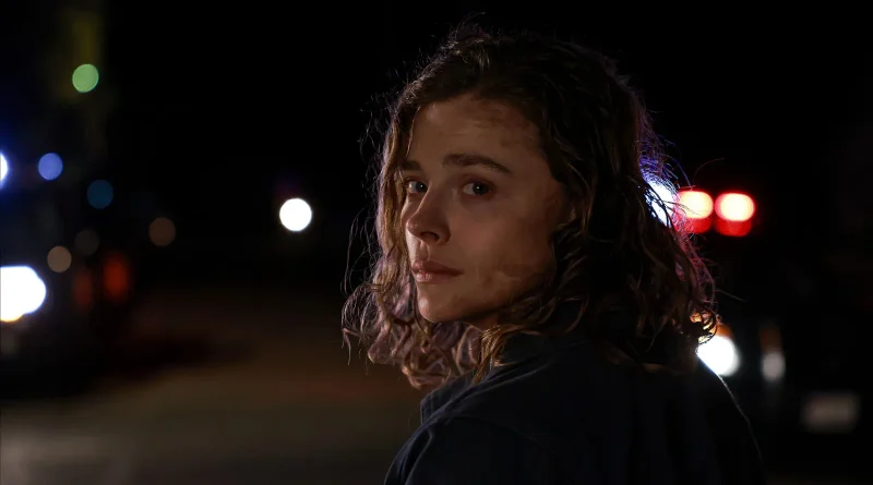 Primer plano de Chloë Grace Moretz mirando intensamente por encima del hombro en una escena nocturna con luces de policía desenfocadas de la película Found Alive.
