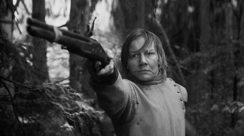 Sandra Hüller apuntando firmemente con una antigua pistola hacia la cámara en un bosque, en una escena en blanco y negro de Rose.