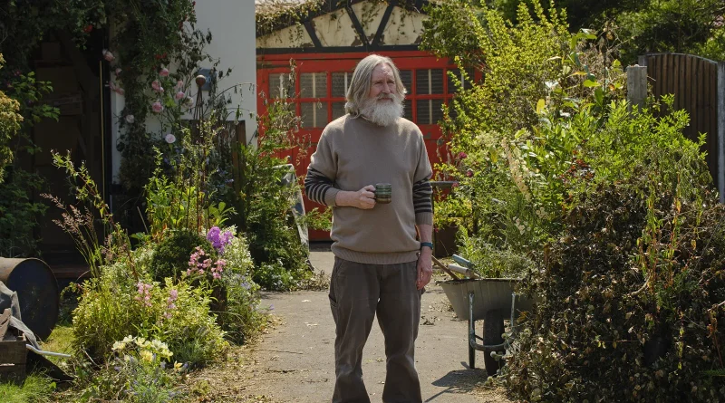 Hombre mayor con barba blanca sosteniendo una taza en un jardín frondoso, en una escena de la serie Pequeños profetas.