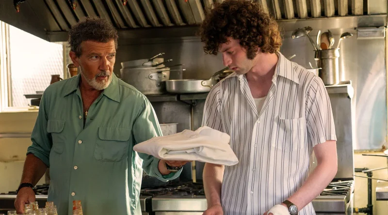 Texto alternativo (alt): Antonio Banderas ofreciendo una chaqueta blanca doblada a un joven de pelo rizado en una cocina industrial en la película Tony.